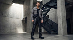 A man stands in a concrete industrial interior wearing a gray utility jacket over a black shirt and black leather pants with black boots.