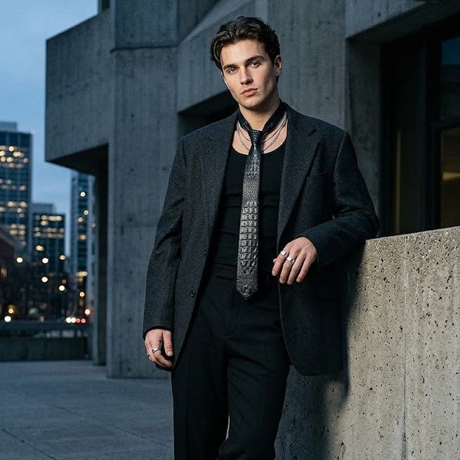 A man wearing a dark charcoal gray suit, black shirt, and a patterned gray tie leans against a concrete wall in an urban setting at dusk.