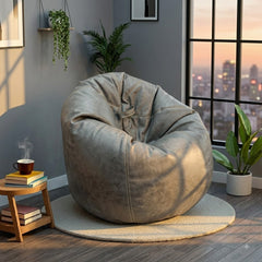A large, plush bean bag chair in a weathered taupe-gray color, styled on a round light beige rug in a modern room with a window showing a city skyline at sunset.