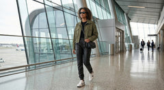 A woman walking through an airport terminal wearing an olive green utility jacket, black t-shirt, black jeans, white sneakers, and carrying a black shoulder bag.