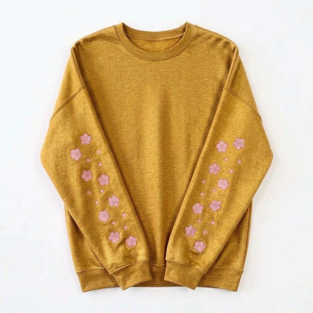 A mustard yellow crewneck sweatshirt with pink embroidered cherry blossom patterns down both sleeves, laid flat on a white background.