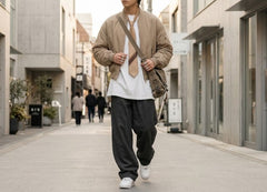 A man walking down a modern concrete alleyway wearing a beige bomber jacket, white t-shirt, dark gray baggy sweatpants, white sneakers, and carrying a brown shoulder bag.