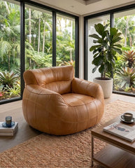 A modern tan leather armchair with a rounded, tufted design, positioned in a bright room with large windows overlooking a lush green garden. A wooden coffee table and a woven rug are in the foreground.