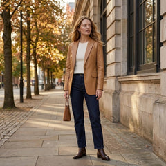 A woman stands on a city sidewalk wearing a tan blazer, white top, dark blue jeans, and brown ankle boots, holding a small tan handbag. Trees with autumn leaves line the street beside a stone building.