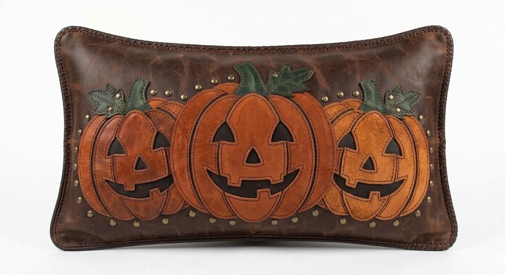 Handmade leather lumbar pillow cover featuring a trio of carved jack-o'-lantern appliques in burnt-orange suede and distressed saddle-brown leather, framed with antique brass stud accents, displayed against a white background.