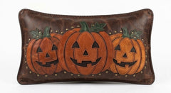 Handmade leather lumbar pillow cover featuring a trio of carved jack-o'-lantern appliques in burnt-orange suede and distressed saddle-brown leather, framed with antique brass stud accents, displayed against a white background.