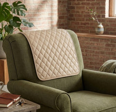 A quilted cream-colored lumbar support pillow resting on the backrest of an olive green armchair in a cozy room with a brick wall background.
