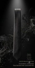 A dark charcoal gray leather tie is shown vertically against a black background with dramatic smoke effects and light speckles. Text at the bottom reads 'SOUTH BEACH LEATHER' and 'ELEGANCE, EVOLVED.