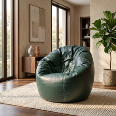 A dark green leather bean bag chair with a drawstring detail, placed on a woven area rug in a modern, sunlit living room with wood floors, a sideboard, and a potted fiddle-leaf fig plant.