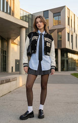 A woman stands outdoors in front of a modern building wearing a varsity-style jacket with black and cream panels and lettering, a light blue shirt, a dark tie, a pleated dark skirt, black tights, white socks, and black loafers.