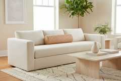 A modern off-white upholstered sofa in a bright, neutral living room setting, styled with a peach quilted lumbar pillow, a light wood coffee table, and a patterned rug.