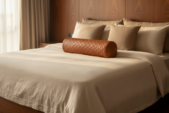 A bed dressed in beige linens with multiple beige pillows and a brown quilted cylindrical bolster pillow, set against a dark wood headboard in a softly lit bedroom.