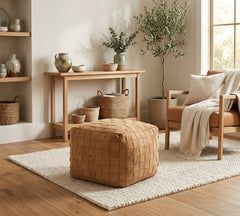 A tan woven leather pouf ottoman sits on a white textured area rug in a bright, modern living room with a wooden console table, a leather armchair, and a potted olive tree.