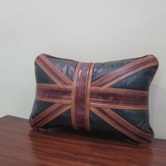 A rectangular throw pillow featuring a Union Jack flag design with dark green and brown faux leather panels, resting on a wooden table against a plain wall.