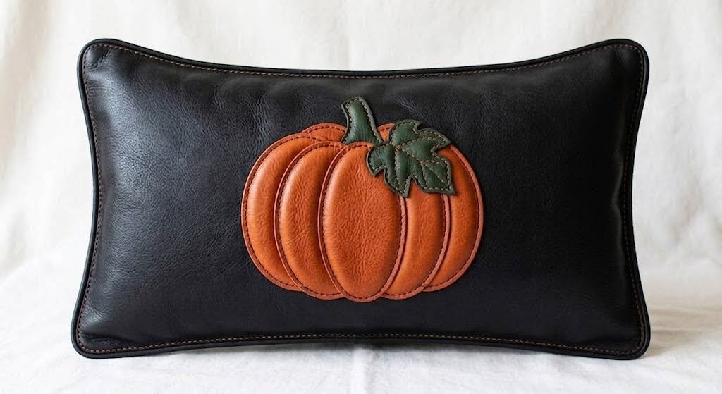 A black leather rectangular lumbar pillow featuring a raised orange leather pumpkin applique with a green leafy stem on the front, displayed against a white fabric background.