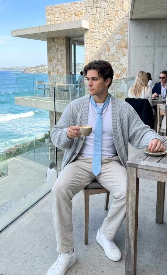 A man sitting on a wooden chair at an outdoor cafe table, wearing a light gray knit cardigan, a light blue tie, a white t-shirt, beige trousers, and white sneakers. The background shows a stone building, glass railing, and the blue ocean.
