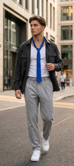 A man walking on a city street wearing a black jacket, white t-shirt, bright blue necktie, light gray trousers, and white sneakers.