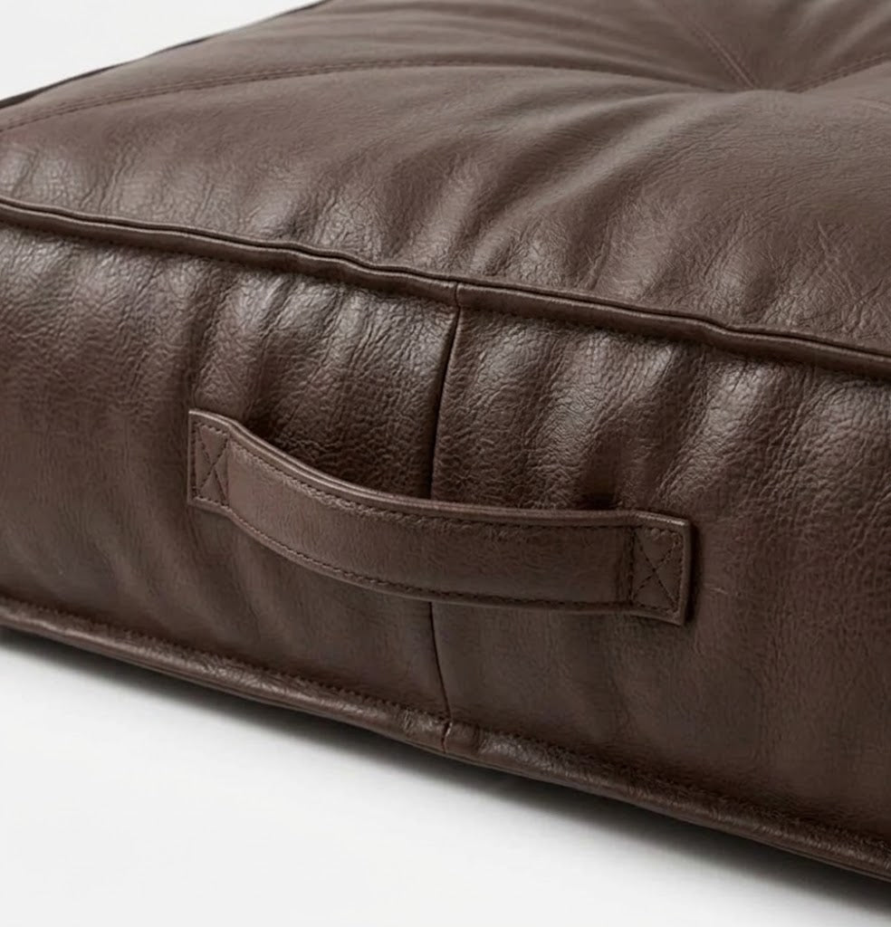 Close-up of a dark brown -leather cushion with a built-in handle, showing the textured leather grain and stitching details.