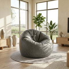 A large, plush gray bean bag chair with a bow detail on the back, set on a round light gray rug in a bright, modern living room with wooden floors, plants, and large windows.