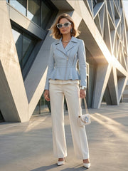 A woman stands outdoors in front of a modern geometric building, wearing a light blue peplum moto jacket, cream wide-leg trousers, white pointed heels, and carrying a small quilted cream handbag.