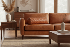 A warm brown leather sofa in a styled living room, featuring a matching quilted cylindrical lumbar pillow, a wooden side table with a vase of pampas grass, and a coffee table in the foreground.