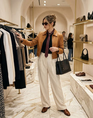 A woman stands in a boutique wearing a tan cropped leather jacket, cream wide-leg trousers, a dark burgundy top, and a dark purple tie. She is holding a black leather tote bag and looking at clothing on a rack.
