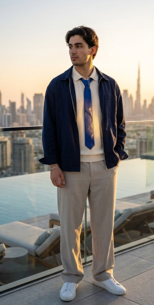 A man stands on a rooftop terrace at sunset wearing a navy blue zip-up jacket over a cream shirt, a blue patterned tie, beige trousers, and white sneakers, with a city skyline in the background.