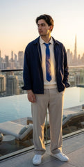 A man stands on a rooftop terrace at sunset wearing a navy blue zip-up jacket over a cream shirt, a blue patterned tie, beige trousers, and white sneakers, with a city skyline in the background.