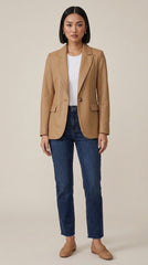 A woman stands against a plain beige studio background wearing a camel tan blazer over a white top, dark blue jeans, and tan loafers.