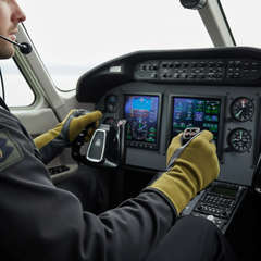 A close-up lifestyle shot of a pilot in a cockpit, wearing a dark gray uniform and mustard yellow gloves while holding the yoke controls. The background shows the instrument panel of the aircraft with digital screens and analog gauges.