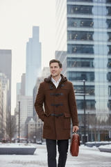 A man wearing a brown duffle coat over a white collared shirt and dark trousers, standing in a snowy city plaza with modern glass skyscrapers in the background and holding a brown leather briefcase.