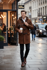 A man walking on a city street wearing a brown wool duffle coat over a white shirt and black trousers, holding a takeaway coffee cup.