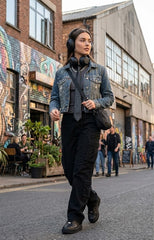 A woman walking down an urban street wearing a dark washed denim jacket, black cargo pants, black boots, and black over-ear headphones around her neck, with a black shoulder bag.