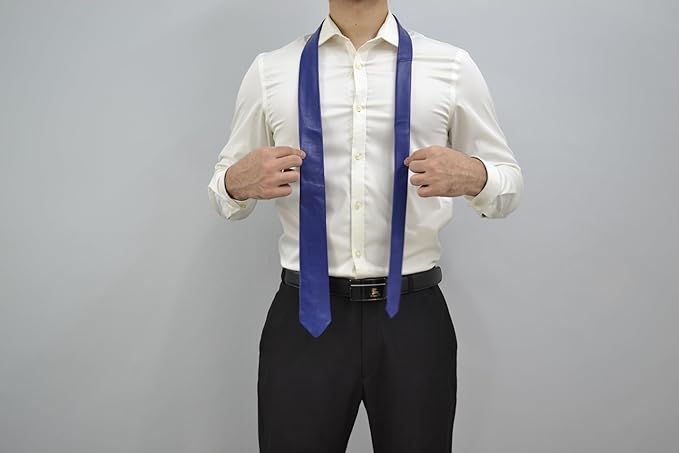 A man wearing a white dress shirt and black trousers models a royal blue necktie, holding the sides of the tie to show its shape and width against a plain gray background.