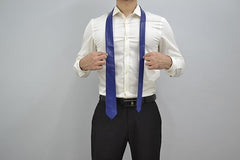 A man wearing a white dress shirt and black trousers models a royal blue necktie, holding the sides of the tie to show its shape and width against a plain gray background.