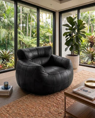 A modern black leather club chair with a tufted quilted design, placed in a stylish living room setting with large windows overlooking a lush tropical garden.
