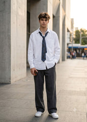 A young man standing outdoors in an urban setting, wearing a white long-sleeve button-up shirt, a loosely knotted dark blue tie, dark charcoal gray trousers, and white sneakers.