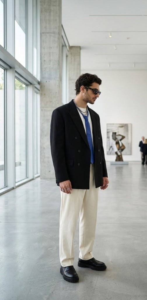 A man wearing a black double-breasted blazer, cream trousers, a blue shirt, and black loafers stands in a modern, sunlit art gallery.