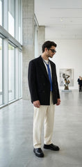 A man wearing a black double-breasted blazer, cream trousers, a blue shirt, and black loafers stands in a modern, sunlit art gallery.