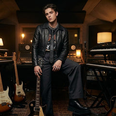 A man wearing a black leather jacket, dark shirt, and wide-leg jeans stands in a music studio surrounded by electric guitars, keyboards, and musical equipment.