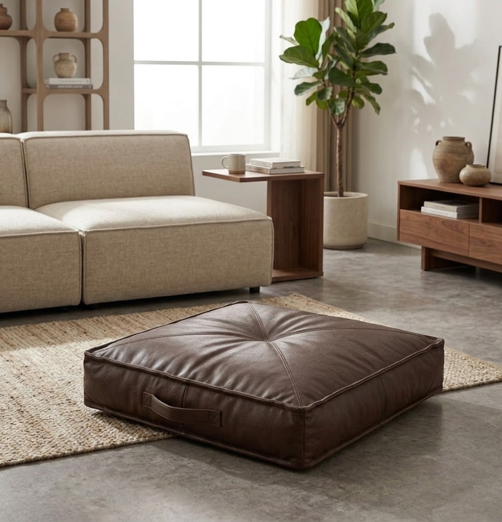 A square brown leather floor cushion with a tufted center and side handle, placed on a woven rug in a modern living room setting.