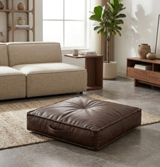 A square brown leather floor cushion with a tufted center and side handle, placed on a woven rug in a modern living room setting.