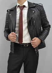 A man wearing a black leather moto jacket over a white dress shirt and a dark red tie, paired with black trousers and a black belt, against a plain gray background.