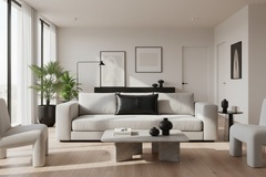 A modern living room featuring a light beige sofa with a black accent pillow, a low concrete coffee table, and a large potted plant near tall windows with sheer curtains.