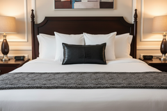 A neatly made bed in a hotel-style room, featuring white linens, a dark wood headboard, a textured dark gray throw blanket, a black textured accent pillow, white pillows, and matching bedside lamps.
