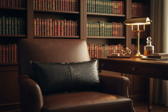 A brown leather armchair with a black crocodile-embossed lumbar pillow, set in a classic library with wooden bookshelves and a desk.