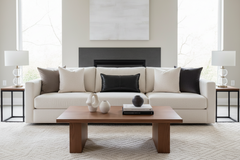 A modern living room setup featuring a cream-colored sectional sofa with black and taupe accent pillows, a wooden coffee table, and matching side tables with white table lamps.