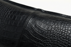 Close-up of a black crocodile-embossed leather surface with visible grain texture and stitching, photographed against a white background.