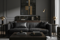 A dark leather sofa in a moody, modern living room setting with a dark wood coffee table, gold table lamps, a marble console, and a large abstract art piece on the wall.