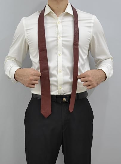 A man wearing a white long-sleeve dress shirt and black trousers, with a dark brown tie draped around his neck like a neck strap, against a plain gray background.